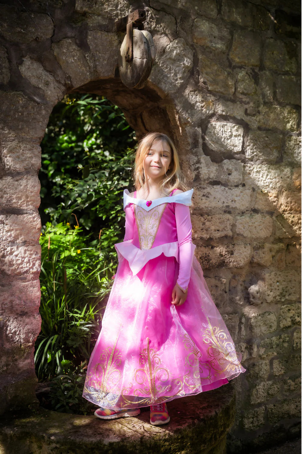 Girl in sleeping beauty dress in front of brick archway. Pretty gold swirls and designs cover the bottom and center of the pink dress.