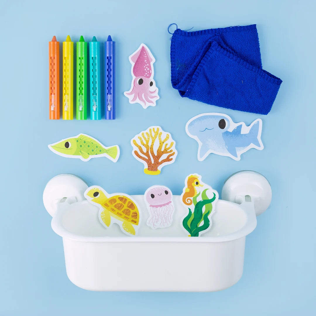 A children's bath toy set arranged on a light blue background, including a white tub with suction cups, colorful crayons, a blue cloth, and sea-themed stickers or toys such as a squid, fish, coral, shark, turtle, jellyfish, and seahorse.