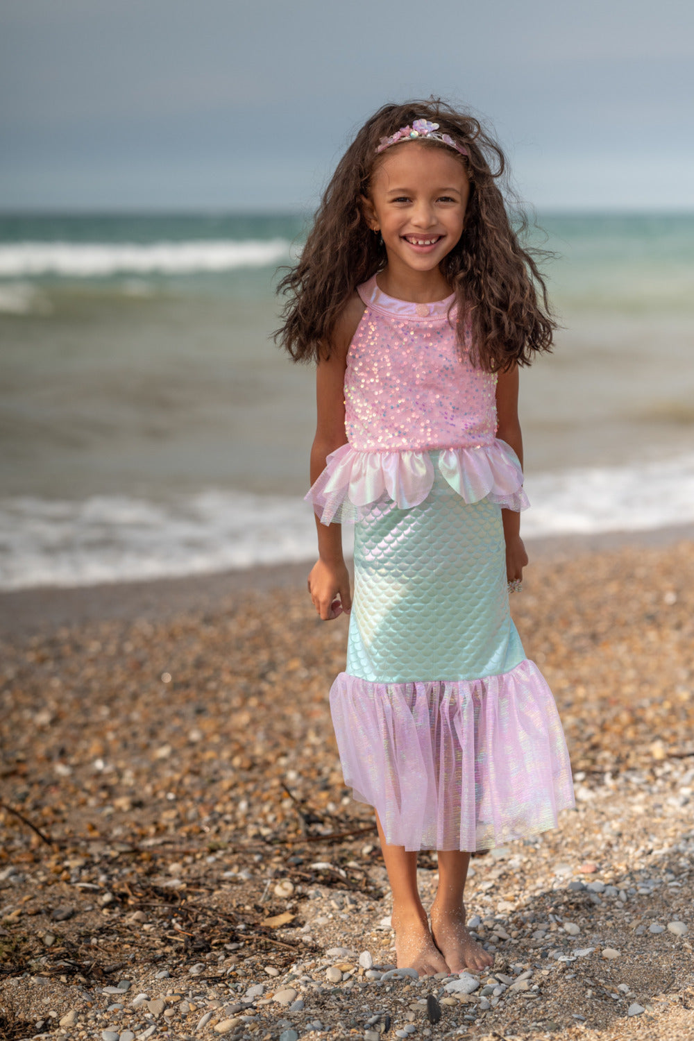 Young girl in a mermaid-themed dress standing on a beach with ocean waves in the background.