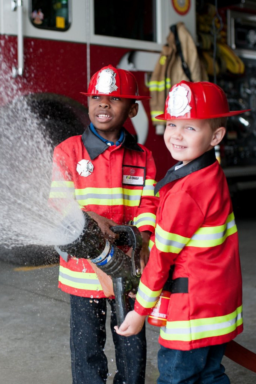Two children dressed in red firefighter outfits with reflective tape details, playing with a water gun simulated as a fire extinguisher.