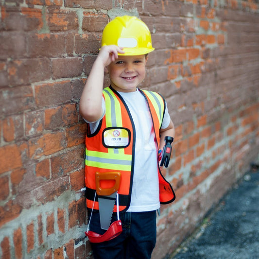 A child dressed in a construction worker costume with reflective orange vest, hard hat, and tools standing against a brick wall.