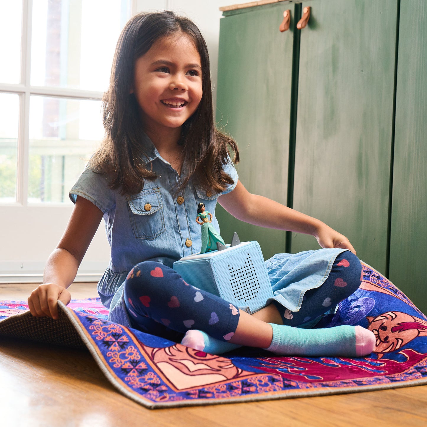 Happy young girl sits cross-legged on a patterned rug, playing with a light blue toniebox that has a small jasmine figurine on top.
