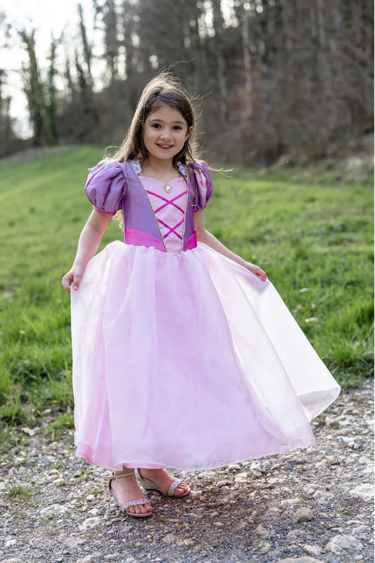 Young girl in a pink and purple Rapunzel inspired dress standing outdoors on a gravel path with grass and trees in the background.