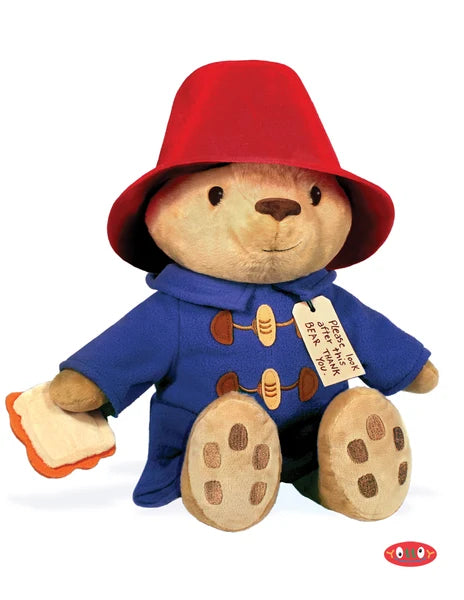 A plush teddy bear dressed as Paddington Bear, wearing a red hat and blue duffle coat, seated against a plain white background with a small red and white logo in the bottom right corner.