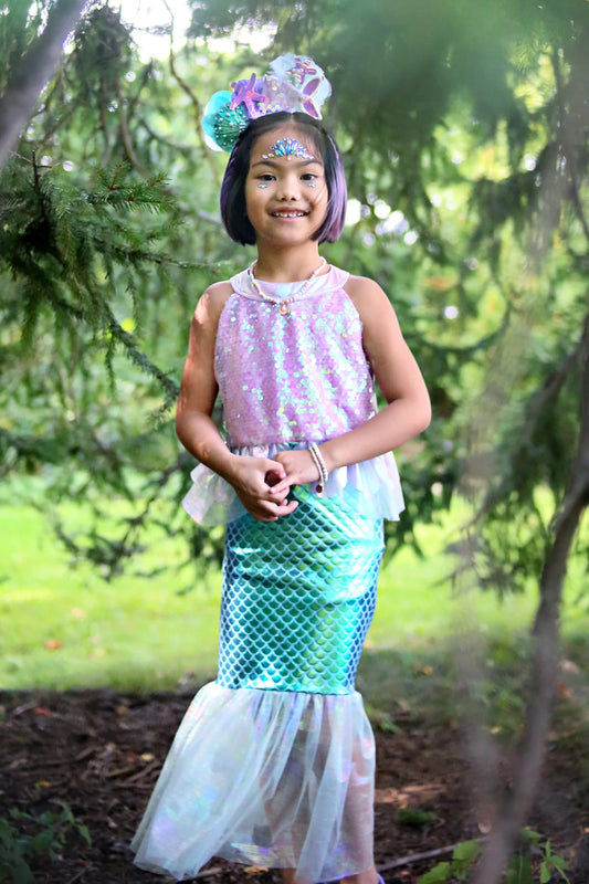 A child wearing a Misty Mermaid Dress with a pink high-neck top and metallic teal skirt, with a mermaid-style skirt made of tulle, standing in a garden setting.