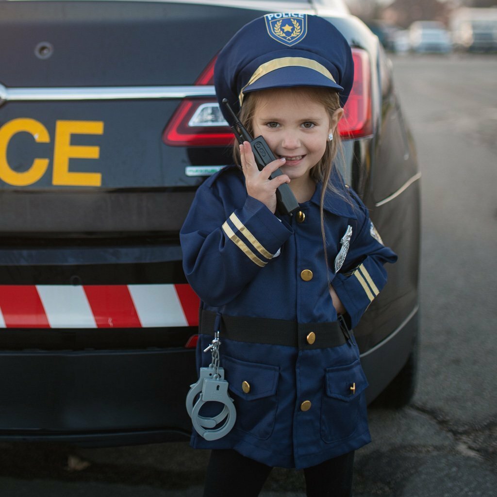 A child dressed in a police officer costume, complete with a jacket, hat, handcuffs, and a walkie talkie, standing next to a police car.