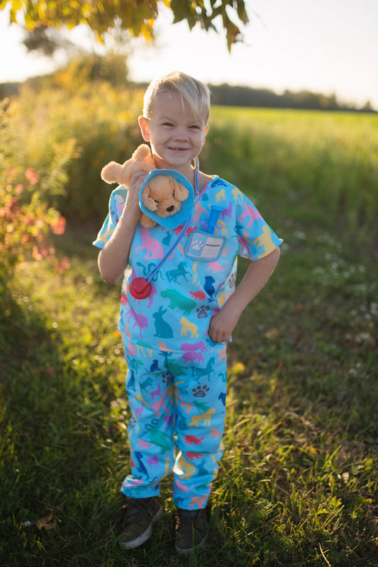 A child dressed in blue veterinarian scrubs with a multicolored pattern, holding a plush toy and wearing a toy stethoscope.