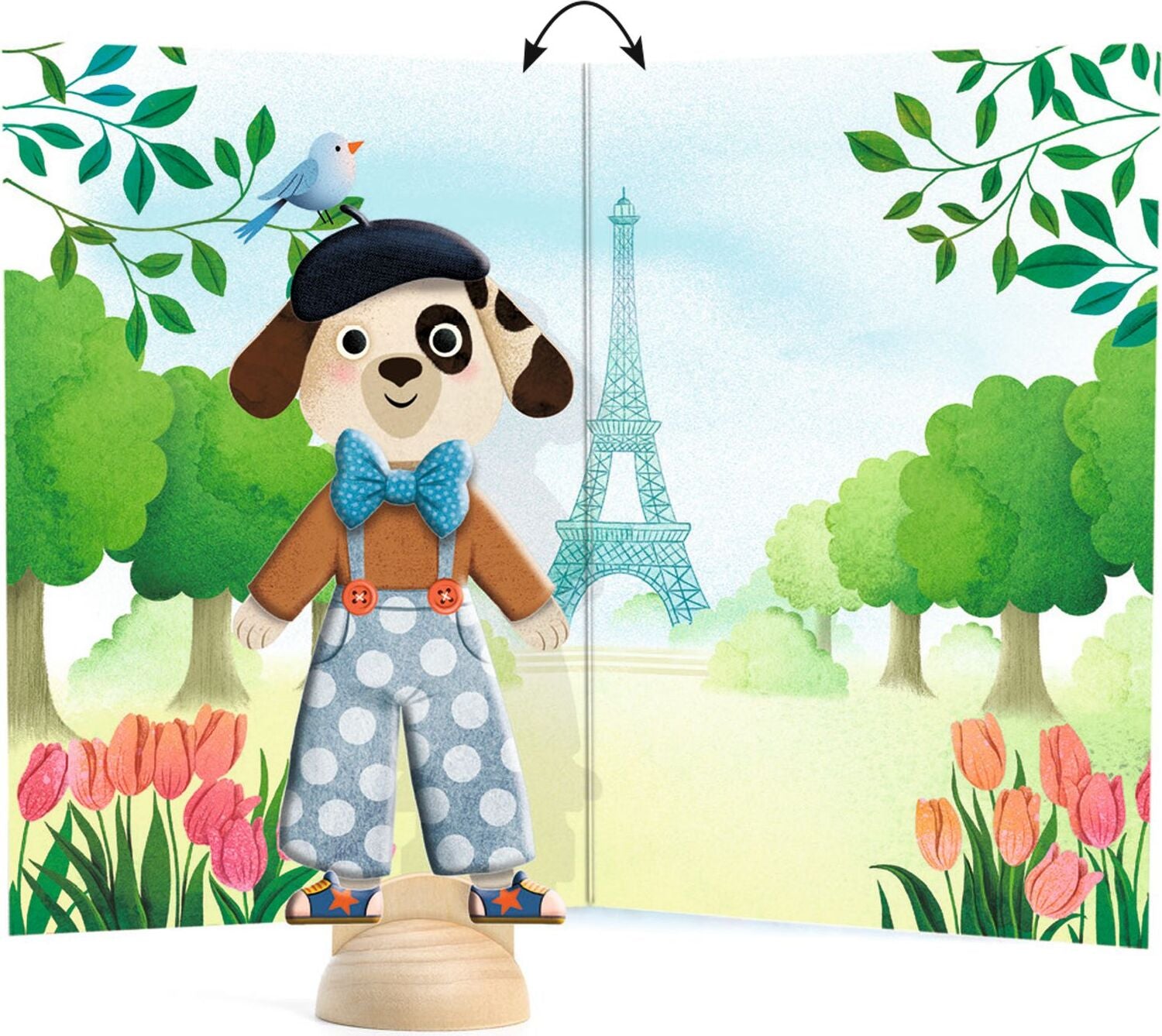 A wooden dog doll on a rotating base, shown in front of a printed backdrop depicting a Parisian street scene with the Eiffel Tower, trees, tulips, and a bird. A black arrow graphic indicates the base can rotate.