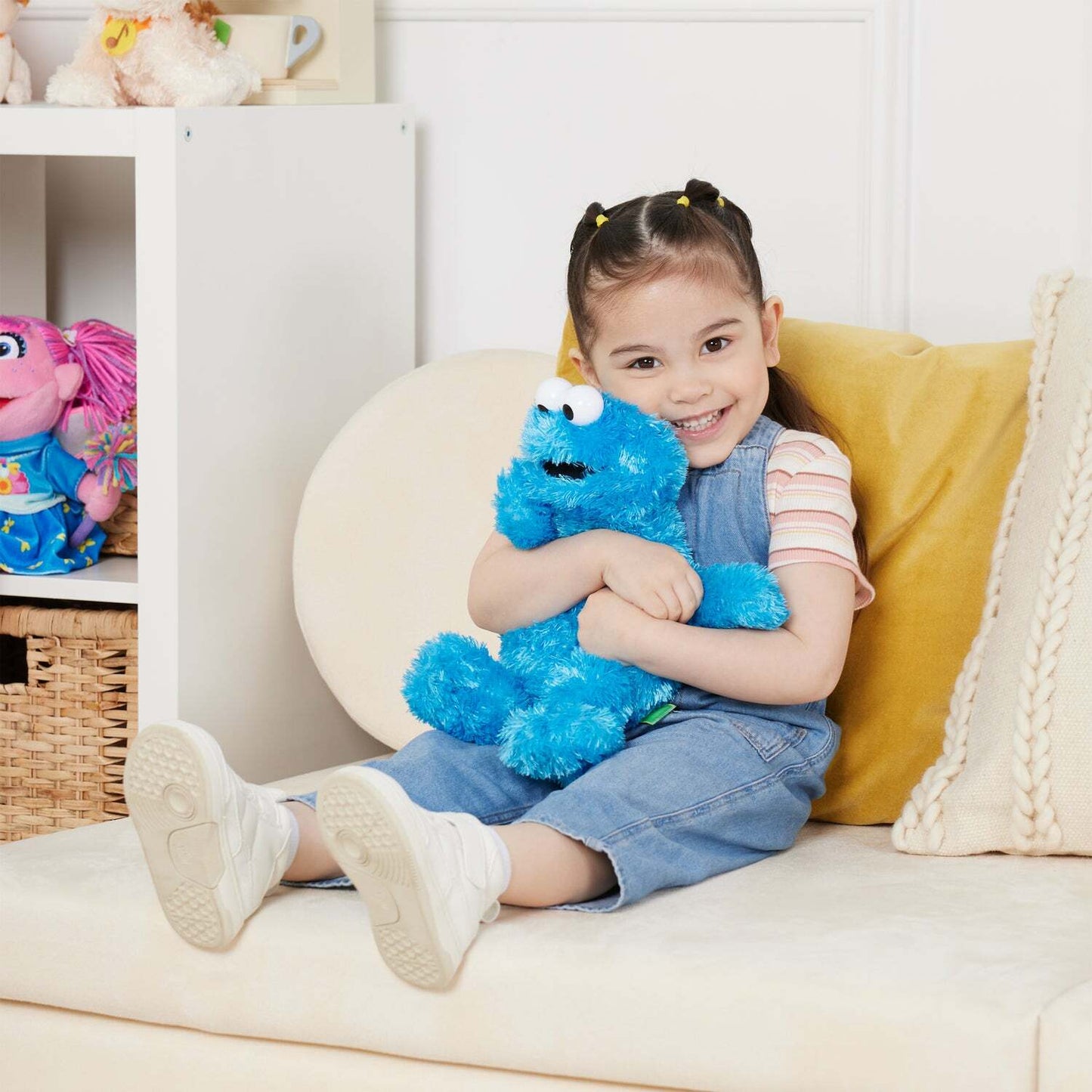 Child sitting on a couch holding a blue plush toy, with a white shelf and colorful toys in the background.