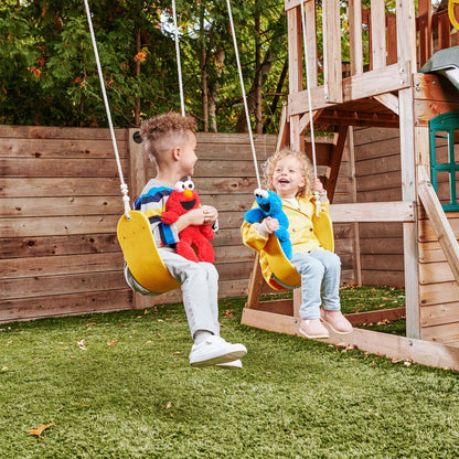 This image shows two young children playing together  on a wooden swing.
On the left, a boy sitting on a yellow swing holds a red Elmo plush toy. On the right, a young girl sitting on an adjacent yellow swing holds a grover  plush toy while laughing happily. 