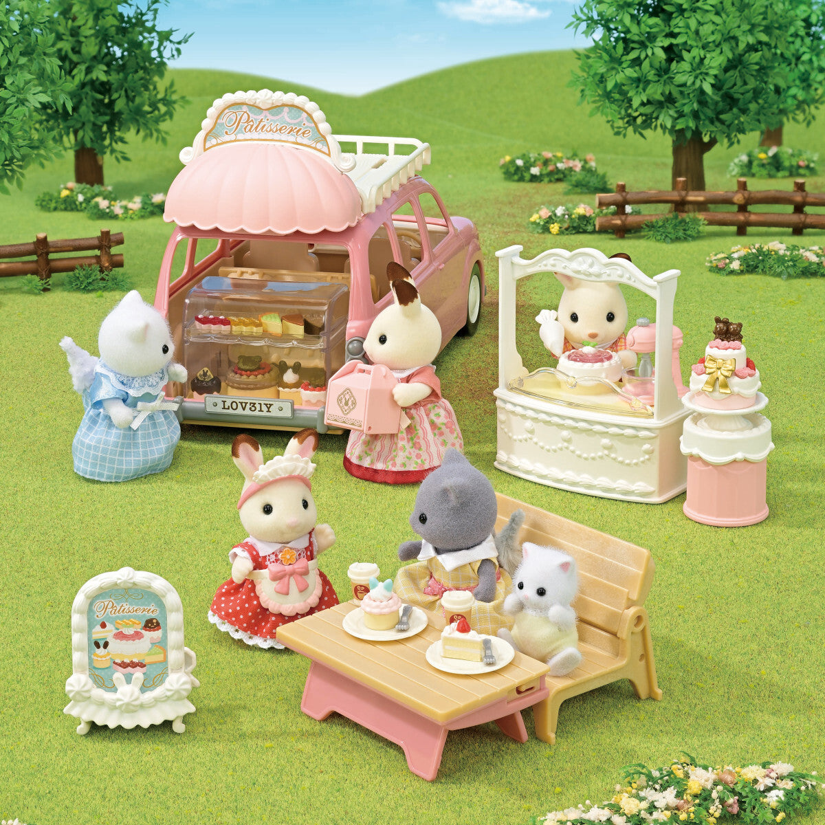 A outdoor play scene featuring small calico critters animal figures enjoying a pink bakery shop van, with a display counter, cake stand, and a table set with miniature pastries on a green grassy lawn.