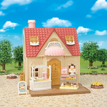 A calico critters cream and pink toy bakery building with a red tiled roof set against an outdoor park background, featuring a front counter, display case filled with miniature cakes, and an entryway sign.