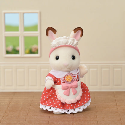 A small, white CALICO CRITTER rabbit figure standing in a room, wearing a red polka-dot dress, a pink apron with a floral motif, and a white baker's hat.