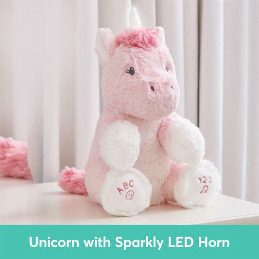 pink plush unicorn sits upright against a light background, Her right foot features embroidered pink text that reads "ABC" with a magnifying glass icon, while her left foot displays "123" with musical notes.
a teal banner states "Unicorn with Sparkly LED Horn" in white text.
