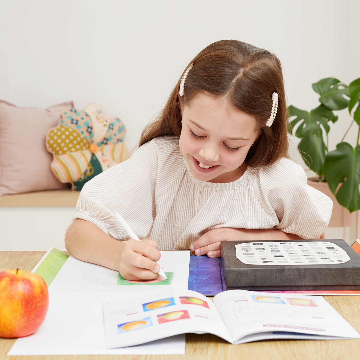 Young girl writing in a notebook with an apple and coloring book on a table.