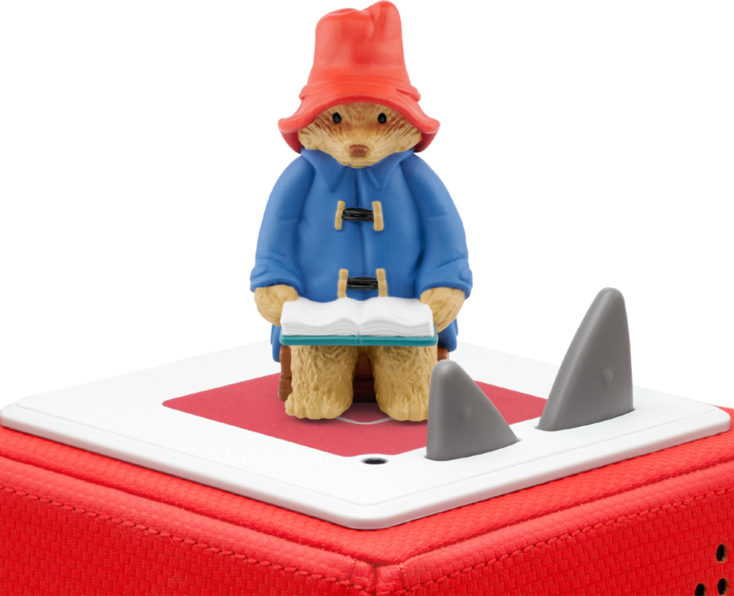 A figurine of Paddington Bear wearing his blue coat and red hat, holding a book on a top of a red toniebox.