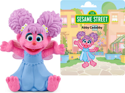 Abby Cadabby figurine with pink hair and blue outfit, sitting with arms open, alongside a Tonie packaging featuring Sesame Street branding and an illustration of Abby Cadabby.