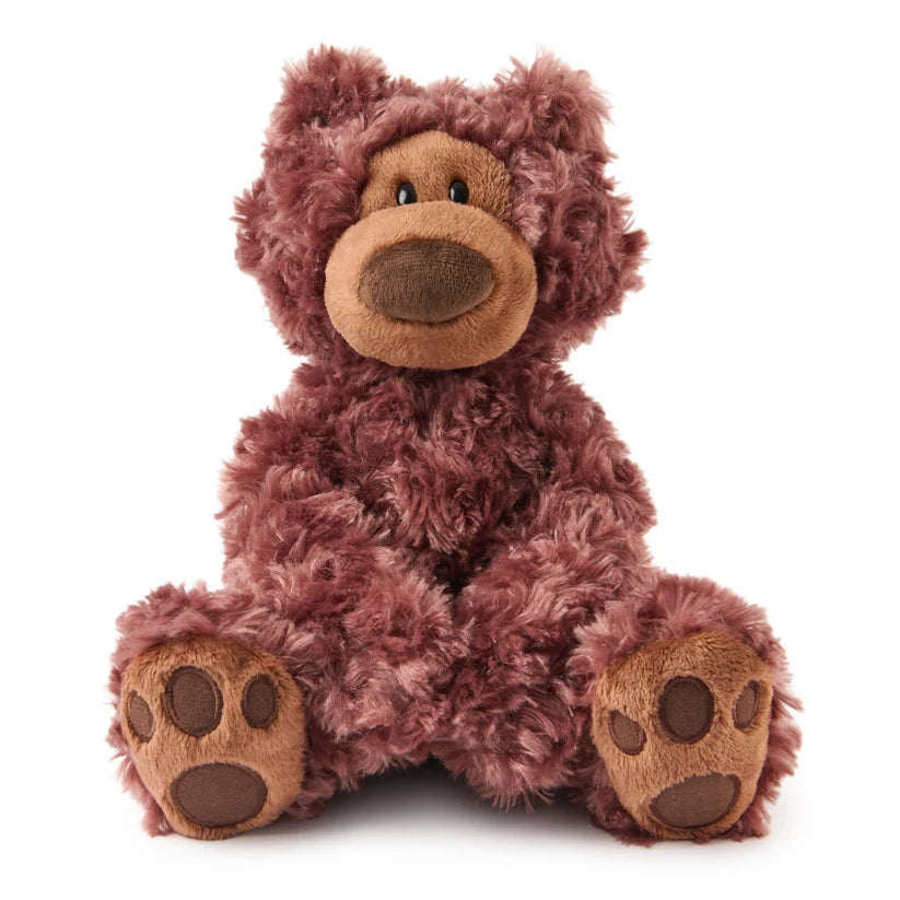 A plush, shaggy teddy bear with deep plum-purple fur sits facing forward on a plain white background. It features a light tan muzzle with a dark brown oval nose, small black bead eyes, and smooth tan paw pads on its feet.
